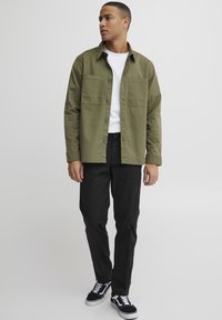 Olive green button-up shirt with two chest pockets, worn over a white t-shirt, paired with black trousers and black sneakers.