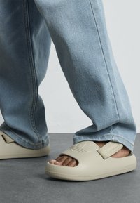 Bežna natikača z odprto konico in teksturiranim trakom, nosena z svetloblue jeans hlačami. Gladek material in minimalističen dizajn.