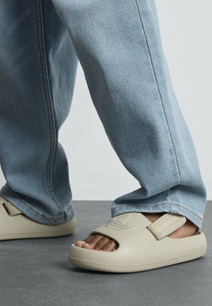 Bézs színű, nyitott orrú strandpapucs textúrázott pánttal, világoskék farmernadrággal viselve. Simító anyag és minimalista dizájn.