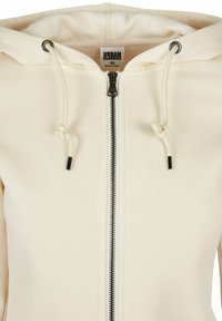 Gräddfärgad zip-up hoodie med en dragsko på huven, prydd med en svart dragkedja och detaljer på dragsnörena. Tyget verkar mjukt och strukturerat.