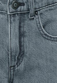 Tuvo plāna skatiena uz pelēkām denim biksēm, kuras demonstrē pogu, jostu cilpu, priekšējo kabatu un redzamās šuves detaļas uz auduma tekstūras.