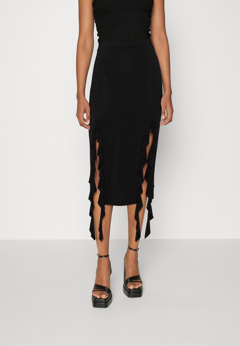 Femme portant une jupe midi noire avec des fentes latérales froncées, associée à des sandales noires à plateforme et bride à la cheville.