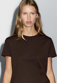 Brun bomulds-T-shirt med korte ærmer og rund halsudskæring, som har en glat tekstur og et enkelt, minimalistisk design.