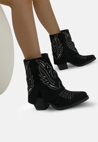 Piernas que llevan botas vaqueras de ante negro con patrones de llamas bordadas en blanco y tacones bajos, sentadas en una silla blanca.