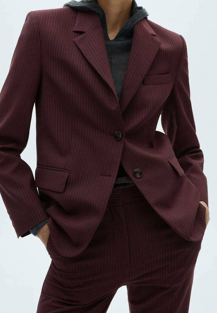 Personne portant un costume bordeaux à fines rayures avec un blazer deux boutons et un pantalon assorti, superposé à un sweat à capuche gris foncé.