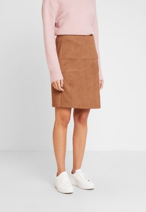 Persona con falda marrón de ante hasta la rodilla, suéter de manga larga rosa y zapatillas blancas, de pie contra un fondo liso.