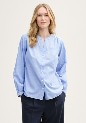 Chemisier bleu clair à col mandarin, orné de petits motifs géométriques blancs, avec des manches longues et des poignets froncés.