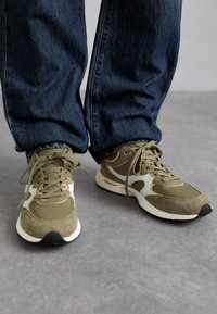 Oliven grønne sneakers med ruskind og mesh-materialer, hvide accenter, guld detaljer omkring snørebåndene og en tekstureret grå sål.
