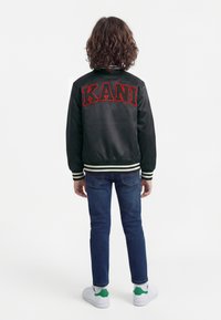 Dítě se středně dlouhými kudrnatými vlasy, oblečené v černé varsity bundě s červeným nápisem „KANI“, modré džíny a bílé tenisky, stojí zády.