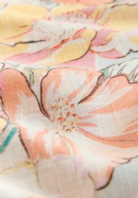 Tissu léger avec un grand imprimé floral rose, jaune et orange et une texture tissée visible, légèrement froissé.