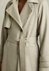 Beige trenchcoat i texturerad tyg med stor krage, knappstängning, midjebälte och sidofickor.