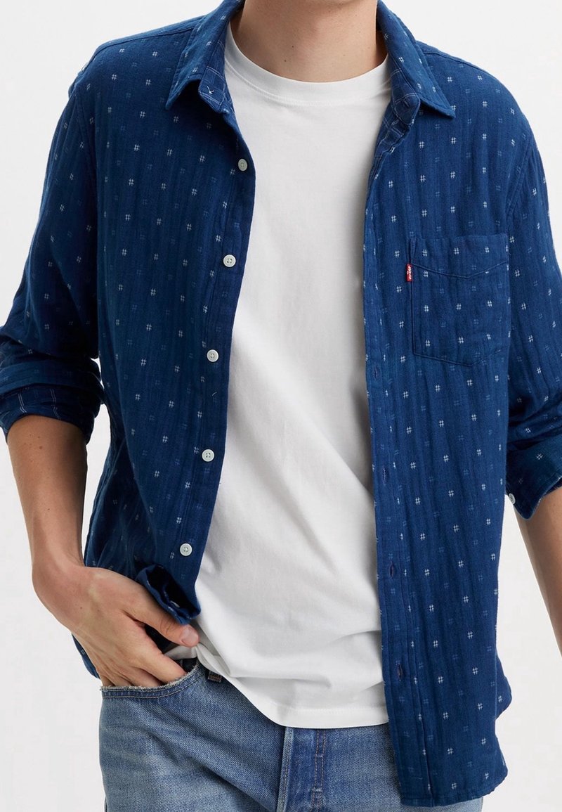 Camicia a quadri blu con dettagli bianchi, realizzata in un tessuto leggero. Presenta una tasca e un colletto casual, indossata sopra una t-shirt bianca.
