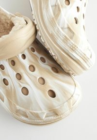 Sabots beiges avec une surface texturée, présentant des trous de ventilation circulaires et un doublure douce. Le design inclut un léger marbrage blanc.