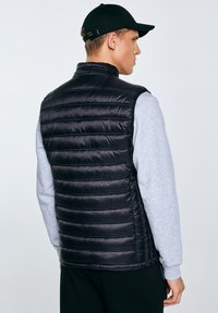 Man draagt een zwart doorgestikt vest over een grijze trui en een zwarte pet, kijkt opzij tegen een effen achtergrond.