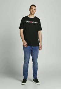 Homem de pé, vestindo uma t-shirt preta "JACK & JONES", jeans azuis e ténis pretos, contra um fundo cinza claro liso.