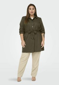 Olivově zelený trenčkot s ohrnutými rukávy, na knoflíky a s páskem v pase, v kombinaci s béžovými kalhotami širokého střihu a sandály s otevřenou špičkou.