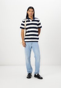 Camisa polo riscada em azul-marinho e branco, feita de tecido texturizado, combinada com jeans azul-claro e sapatos pretos. O modelo segura uma bolsa em creme.