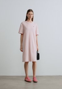 Ροζ βαμβακερό φόρεμα με κοντά μανίκια και άνετη εφαρμογή, με μικρό λογότυπο στο στήθος. Συνδυάζεται με ροζ αθλητικά παπούτσια και μαύρη τσάντα χειρός.