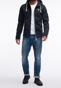 Granatowa bluza zapinana na zamek, wykonana z textured materiału, z troczkami i małym logo. Połączona ze jasnoniebieskimi dżinsami i czarnymi trampkami.