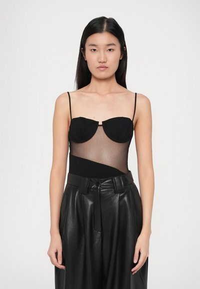 Une jeune femme aux longs cheveux bruns porte un body noir transparent avec des bonnets structurés et un pantalon en cuir noir taille haute, faisant face.
