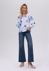 Blusa floreale bianca con dettagli blu, polsini elastici e cappuccio. Abbinata a jeans svasati blu scuro e sandali con tacco bianchi. Sfondo semplice.