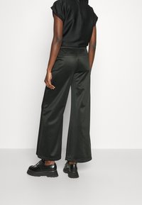 Mujer vestida con pantalones de pierna ancha negros, top negro de manga corta y zapatos de plataforma negros, de pie contra un fondo blanco liso.