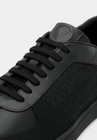 Μαύρο δερμάτινο αθλητικό παπούτσι με μαύρα κορδόνια, ανάγλυφο λογότυπο στη γλώσσα, και υφασμάτινο πλευρικό πάνελ με διακριτικό επαναλαμβανόμενο μοτίβο.