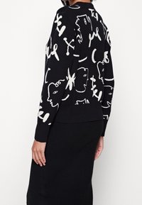 Schwarzer Pullover mit weißen abstrakten Gesichtern und Textmustern. Hat lange Ärmel, einen runden Halsausschnitt und gerippte Bündchen.