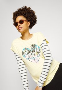 T-shirt amarelo claro com um gráfico de um coração, palmeiras e ondas. Camada de uma camisa de manga longa às riscas. Óculos de sol castanhos.