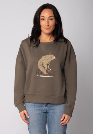 Vrouw met lang donker haar in een bruine sweatshirt met een illustratie van een beer die op een rode fiets rijdt, gecombineerd met lichtblauwe jeans.
