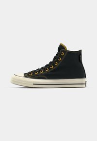 Las zapatillas altas negras cuentan con una parte superior de lona, costuras amarillas, ojales dorados y una suela de goma blanca en contraste con franjas negras.