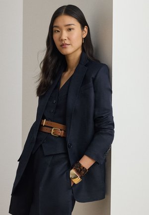 Ženska z dolgimi temnimi lasmi nosi temnomodro suknjič in telovnik, rjavi dvojni pas ter masivne zlate in želvje lupine zapestnice, stoji ob steni.