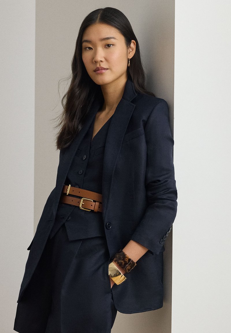 Γυναίκα με μακριά σκούρα μαλλιά φοράει μπλε σκούρο μπλέιζερ και γιλέκο, καφέ ζώνη διπλής στερέωσης και χοντρά χρυσά και καμηλοπάρδαλη βραχιόλια, στέκεται δίπλα σε έναν τοίχο.