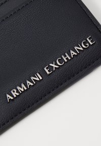 Mørkeblå lærpung med preget tekst "ARMANI EXCHANGE." Har en strukturert overflate og rene kanter. Inkluderer kortlommer.