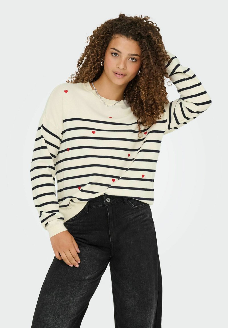 Gestreepte trui in crème met zwarte en rode hartaccenten; ronde halslijn; lange mouwen; gecombineerd met hoge taille zwarte jeans.