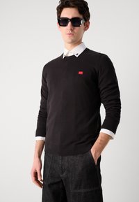 Pulover negru tricotat cu gât în V, având un mic logo roșu. Purtați peste o cămașă albă cu guler, asortat cu blugi închiși la culoare și ochelari de soare.