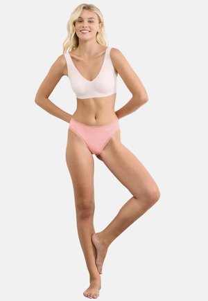 Smiling woman with long blonde hair stands barefoot, wearing a light beige bra and pink lace-trimmed underwear against a plain white background.