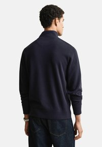 Maglione blu navy scuro con collo alto, orlo e polsini a costine, tessuto liscio e un dettaglio sottile di cucitura sul retro. Indossato con jeans scuri.
