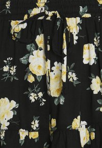 Černá tkanina s žlutým květinovým vzorem, se stahovacími šňůrkami v pase a nabranými detaily. Materiál vypadá lehce a texturovaně.