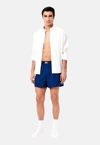Chemise blanche à boutons, short bleu marine avec une taille élastique, et chaussettes blanches. La chemise a une coupe décontractée, un ourlet droit et un détail de poche.