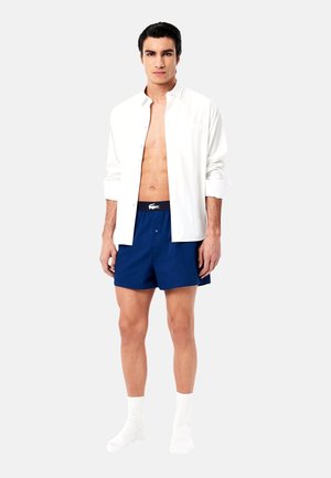 Chemise blanche à boutons, short bleu marine avec une taille élastique, et chaussettes blanches. La chemise a une coupe décontractée, un ourlet droit et un détail de poche.