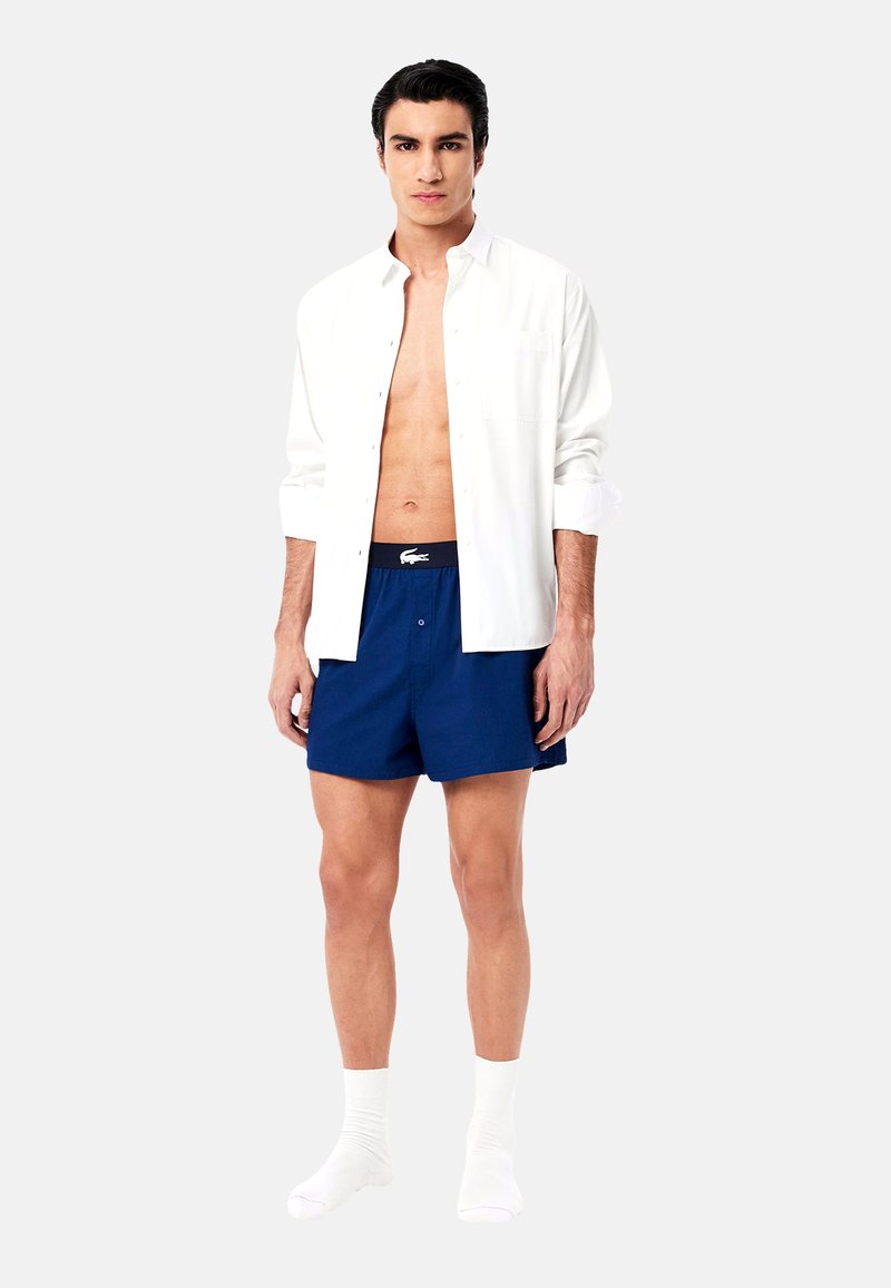 Chemise blanche à boutons, short bleu marine avec une taille élastique, et chaussettes blanches. La chemise a une coupe décontractée, un ourlet droit et un détail de poche.