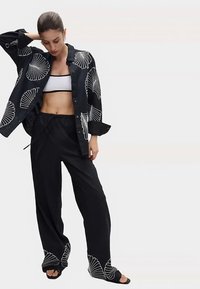 Zwart overhemd met wit schelpenpatroon, gecombineerd met een zwart cropped topje en losse zwarte broek. Gladde stof en ontspannen pasvorm. Zwarte sandalen.