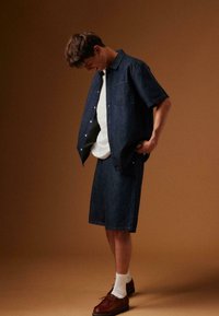 Jeune homme regardant vers le bas, portant une chemise en jean foncé et un short, avec des chaussettes blanches et des chaussures marron, sur un fond marron uni.