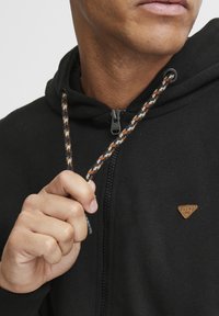 Sudadera negra con cremallera que presenta una tela texturizada, con un cordón trenzado en contraste y un pequeño parche con logo marrón en el pecho.