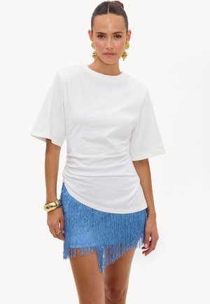 Femme aux cheveux tirés en arrière portant un haut blanc à manches courtes, une jupe bleue à franges, des boucles d’oreilles anneaux dorés et un bracelet épais.