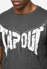 Grijs T-shirt met grote, verweerde witte "TAPOUT" opdruk, ronde halslijn en korte mouwen met een licht vervaagde textuur.