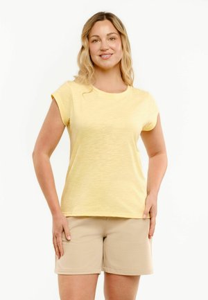 Smilende kvinde med bølget blondt hår iført en lysegul kortærmet skjorte og beige shorts til knæene, stående med hænderne langs siden.