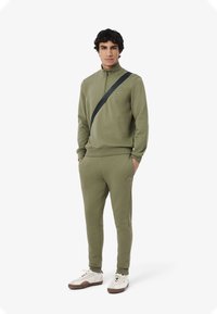 Olijfgroene trainingsuit van zacht materiaal, met een hoge kraag en rits. Bevat een zwarte band over het lichaam en ribbelmanchetten.