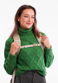 Zelený pletený rolák s diamantovým vzorem, doplněný světlobéžovými popruhy od batohu. Model má na sobě šedé kalhoty a drží popruhy.